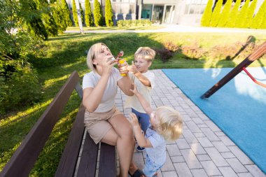 Bir anne, canlı su oyuncaklarıyla oynarken iki küçük oğluyla oyun parkında neşe içinde etkileşim kurar. Çocuklar hevesle uzanıyorlar, eğlenceli aktivitelerinden heyecan duyuyorlar, etrafları onlarla çevrili.