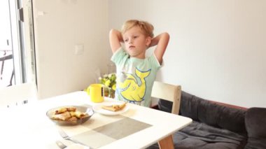 Genç beyaz bir çocuk, iyi aydınlatılmış bir mutfakta lezzetli bir atıştırmalığın tadını çıkarırken rahatlıyor. Bir tabak yemek ve neşeli bir içecekle masada oturuyor, memnun ve rahat görünüyor.