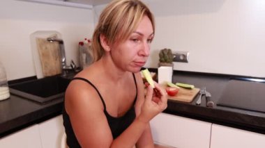 Sıcak bir mutfakta, beyaz bir kadın taze sebzelerden bir ısırık alır, yemek hazırlarken lezzetin tadını çıkarır. Etrafı malzemelerle çevrili, gevşemiş ve yemek pişirme süreciyle meşgul görünüyor..