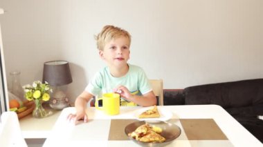 Genç beyaz bir çocuk mutfak masasında oturuyor, parlak sarı bir bardaktan yudumlarken lezzetli yemeğinin tadını çıkarıyor. Mutfak basit ve sıcak. Dekoratif unsurlar da cazibe katıyor.