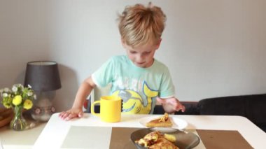 Genç beyaz bir çocuk mutfak masasında oturuyor, bir dilim turtanın tadını çıkarırken sarı bardaktan yudumluyor. Sıcak bir mutfak düzeniyle çevrili, aperatifleriyle meşgul görünüyor..