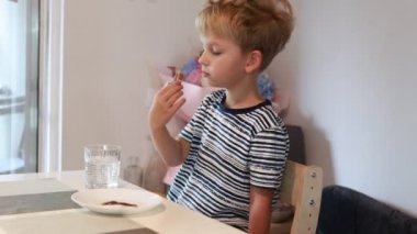 Genç beyaz bir çocuk mutfak masasında oturmuş su içerken aperatifinin tadını çıkarıyor. Parlak ve davetkar bir mutfakta yemeğinin tadını çıkarırken odaklanmış ve memnun görünüyor..