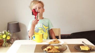 Genç beyaz bir çocuk mutfak masasında oturmuş, heyecanlı bir kahvaltı yapıyor. Kreplerle dolu bir tabağı ve elinde bir fincan var. Mutfak atmosferiyle çevrili..