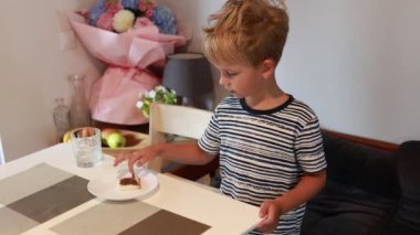 Genç beyaz bir çocuk mutfak masasında oturup lezzetli bir tatlıyı zevkle yiyor. Çiçekler ve meyveler arka planı süsleyerek sıcak ve sıcak bir ortam oluştururken, masa bir tabak içeriyor.