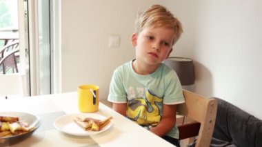 Genç bir çocuk mutfak masasında sessizce kahvaltının tadını çıkarıyor. Yemek dilimlerini barındıran tabağına bakarken düşünceli bir yüz ifadesi var. Yakınlarda renkli bir kupa var.