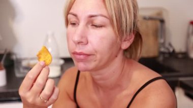 Sarı saçlı bir kadın mutfak tezgahında oturup lezzetli bir atıştırmalığın tadını çıkarıyor. Yemeklere odaklanmış, yemek rutininden zevk aldığını gösteriyor. Mutfağın çağdaş bir yapısı var.