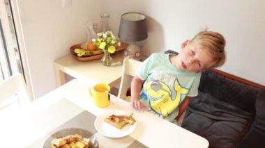 Genç beyaz bir çocuk modern bir mutfakta yemek masasında oturmuş, yemeğin tadını çıkarıyor. Mutfakta atmosfere çiçekler ve meyveler eklenmiş parlak bir dekor yer alıyor. Onun şakacı tavırları,