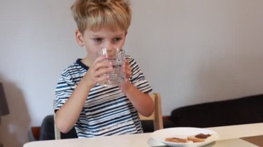 Genç bir çocuk mutfak masasında oturur, aperatiflerinin tadını çıkarırken bardaktan su yudumlar. Mutfakta basit bir dekor ve doğal ışık var. Yemeğini yerken sıcak bir atmosfer yaratıyor..