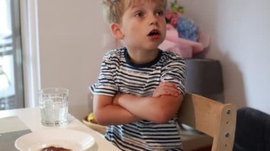 Genç beyaz bir çocuk mutfak masasında kollarını kavuşturmuş, etrafını gözetliyor. İyi ışıklandırılmış ve rahat bir çevreyle çevrili yemeğinden hoşlanıyor gibi görünüyor. Sahne yakalar