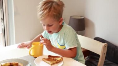 Genç beyaz bir çocuk mutfak masasında oturuyor, sarı bardaktan yudumlarken lezzetli kahvaltı malzemelerinin tadını çıkarıyor. Güneş ışığı odaya dolarken atmosfer sıcak ve davetkar..