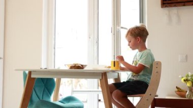 A young Caucasian boy is sitting at a kitchen table, happily eating a nutritious meal. The bright kitchen is filled with natural light, creating a warm atmosphere. He appears focused and content as he