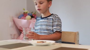 Genç beyaz bir çocuk mutfak masasında oturur, önünde aperatif yerken bardaktan su yudumlayarak mutlu mesut oturur. Atmosfer rahat, dekoratif elementler rahat mutfağı geliştiriyor.