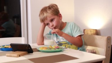 Genç bir çocuk mutfak masasında oturmuş, renkli meyve ve abur cuburlarına dikkatlice bakarken bir içki yudumluyor. Ilık aydınlatma, o keyfini çıkarırken sıcak bir ortam sağlıyor.