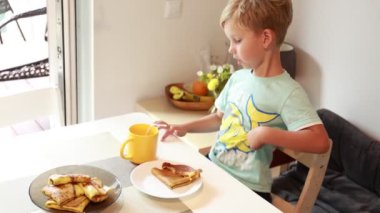 Genç beyaz bir çocuk neşeli bir mutfakta, gözleme kahvaltısı yapmaya hevesle hazırlanıyor. Yakınlarda sıcak bir içecekle önce hangi parçayı yiyeceğine karar verirken odaklanmış görünüyor..