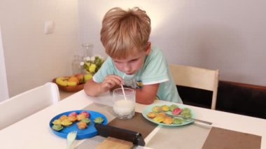 Genç beyaz bir çocuk mutfak masasında oturup renkli abur cuburlar yemeye odaklanmış. O, parlak ikramlar tabaklarla çevriliyken bir bardak sütün tadını çıkarıyor. Mutfakta rahat bir atmosfer var.