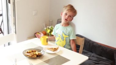 Genç bir çocuk sakince mutfak masasında oturup krep ve meyve suyunun tadını çıkarıyor. Davetkar mutfak güzelce dekore edilmiş, yemeği için rahat bir ortam yaratıyor. Yakınlarda taze meyve sergileniyor.