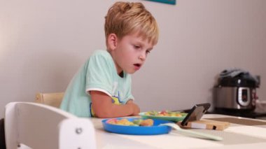 Genç beyaz bir çocuk mutfak masasında oturuyor, önündeki alete odaklanmış. Görünürde ekranda ne varsa onunla uğraşırken iki tabak renkli atıştırmalıkları var..