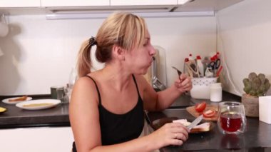 Beyaz bir kadın mutfağında oturmuş lezzetli bir yemeğin tadını çıkarıyor. Taze malzemelerle çevrili bir şekilde içkisini yudumlarken bir lokma yiyecek alıyor. Mutfaktaki atmosfer sıcak bir havayı yansıtıyor.