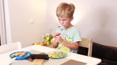Genç bir çocuk mutfak masasında oturup tabağından renkli atıştırmalıklar yiyor. Bir çatal tutuyor ve etrafındaki çeşitli yiyeceklerle yemeğinin tadını çıkarırken mutlu görünüyor..