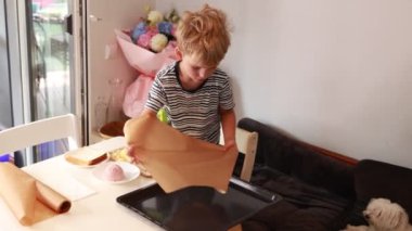 Genç bir çocuk, konforlu bir mutfak ortamında yemek hazırlamakla aktif olarak ilgilenir. Parşömen kağıdını dikkatlice fırın tepsisine yerleştiriyor ve tomurcuklanan aşçılık becerilerini sergiliyor. Bu etkinlik