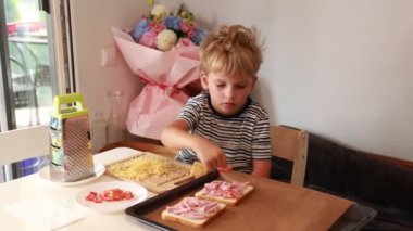 Genç bir çocuk sıcak bir mutfakta hevesle sandviç hazırlıyor. Ekmeğe taze malzemeler ekliyor ve çeşitli yemek aletleriyle eğlenceli ve ilgi çekici bir şekilde etkileşime geçiyor.