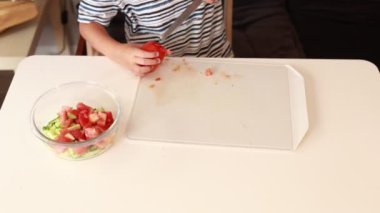 Genç bir çocuk domates doğrayarak ve diğer malzemelerle karıştırarak taze bir salata hazırlıyor. Mutfak seti sıcak ve davetkar. Çocukların yemek pişirme becerilerini gösteriyor.