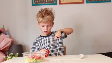 Genç beyaz bir çocuk renkli bir salata yapmak için taze sebzeleri doğrarken dikkatle odaklanıyor. Mutfak dekorasyonu onun yemek pişirme deneyimini arttırıyor, nişanını ve hevesini gösteriyor.