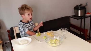 Genç bir çocuk sıcak bir mutfakta taze meyve salatası hazırlıyor. Çeşitli meyveleri dikkatlice dilimliyor, gelişmekte olan aşçılık yeteneklerini sergiliyor ve bu sürecin tadını çıkarıyor..