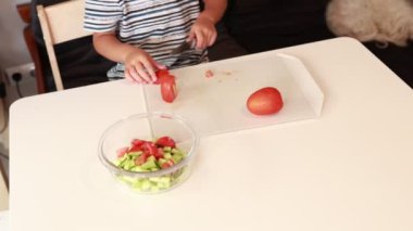 Genç beyaz bir çocuk mutfakta sağlıklı bir yemek hazırlamakla meşgul. Parlak kırmızı bir domatesi ustalıkla doğrarken yanında bir kase doğranmış sebze duruyor.