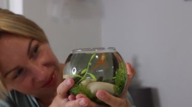 Bir kadın küçük akvaryumuna neşeyle bakıyor, balık ve su bitkileriyle dolu bir cam kaseyi dikkatle tutuyor. Suyun içindeki canlı yaşamdan memnun ve meşgul görünüyor, onu gösteriyor.