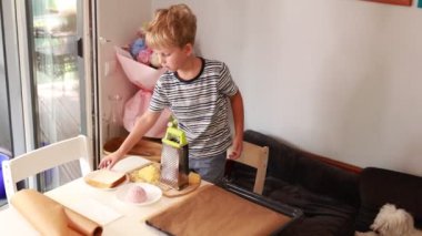 Genç beyaz bir çocuk neşeli bir mutfak ortamında peynir rendelemekle meşgul. Yemek pişirme aletleriyle çevrili bir masada durup lezzetli bir yemek için malzemeleri hazırlıyor. Mutfak.
