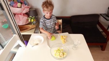 Genç bir çocuk sıcak bir mutfakta taze meyve salatası hazırlıyor. Çeşitli meyveleri ustalıkla kesiyor ve organize ediyor, aşçılık yeteneklerini sergiliyor. Atmosfer rahat ve davetkar.