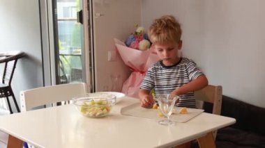 Genç bir çocuk doğal ışıkla dolu parlak bir mutfakta neşeyle taze meyve dilimliyor. Renkli meyve parçalarını özenle düzenlemek için dikkatle işine odaklanıyor. Mutfak neşeli.