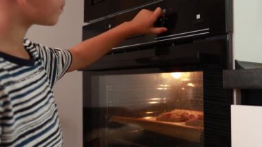 Genç bir çocuk lezzetli ev yapımı pizzası için fırın zamanlayıcısını kurarak yemek pişirmekle meşgul. Yemek pişirme sürecini dikkatle izliyor. Sonuçları görmek için sabırsızlanıyor..