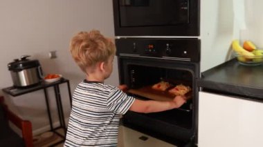 Genç bir çocuk çağdaş bir mutfakta pizza pişirmeye hevesle hazırlanıyor. Fırının önünde duruyor, içindeki pizzaları kontrol ediyor ve yemek pişirme becerilerini keşfediyor. Mutfak çok canlı.