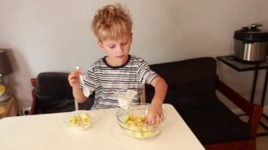 Genç beyaz bir çocuk, sıcacık bir mutfakta taze meyve salatası hazırlıyor. Yemek pişirme deneyiminin tadını çıkarırken bir kasede çeşitli renkli meyveler karıştırıyor. Atmosfer parlak ve...