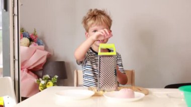 Genç beyaz bir çocuk yemek pişirmeye hevesli, ustalıkla rendeleyici malzemeler üretiyor. Dekorasyon neşeli bir mutfak. Renkli dekorasyonlarla dolu.