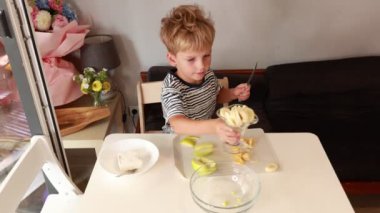 Genç bir çocuk renkli bir meyve salatası hazırlıyor, sıcak bir mutfakta taze malzemeleri dikkatlice doğruyor ve karıştırıyor. Gelişmekte olan yemeklerini sergilerken canlı meyveleri birleştiriyor.