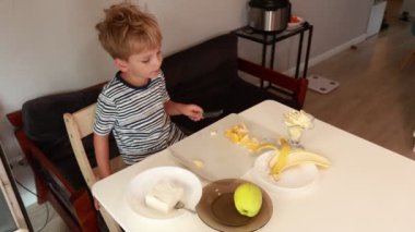 Genç bir çocuk çağdaş bir mutfakta dikkatlice sağlıklı bir aperatif hazırlıyor. O, bir yemek masasında otururken muz ve mango da dahil olmak üzere taze meyve kesiyor. Parlak atmosfer