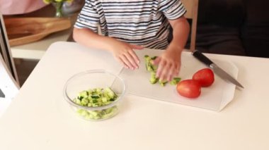 Genç beyaz bir çocuk aktif olarak mutfakta taze sebze salatası hazırlamakla meşgul. Salatalık ve domates doğrarken bu esnada mutfak becerilerini ve yaratıcılığını sergiliyor.