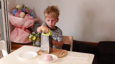 Genç bir çocuk, neşeli bir mutfak ortamında yemek pişirmek için rende kullanıyor. Atmosfer parlak ve davetkar, çiçekler biraz renk katıyor..