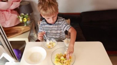 Neşeli bir beyaz çocuk iyi aydınlatılmış bir mutfakta lezzetli bir meyve salatası hazırlıyor. O, yemek pişirme sürecinin tadını çıkarırken çeşitli meyveleri ustaca birleştirir. Atmosfer onun dikkatle izlediği kadar canlı.
