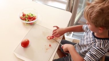 Genç beyaz bir çocuk, parlak bir mutfakta salata için taze malzemeler doğruyor. Domatesleri dikkatlice kesiyor. Yemek pişirme becerilerini ve tutkusunu gösteriyor.