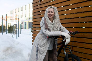 Dışarıda karlı bir ortamda bir bisikletin yanında duran bir kadın, zemini karla kaplı ve arka planda ağaçlar. Kışlık giysiler giyiyor ve bisiklete bakıyor..