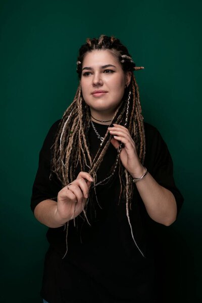 A woman with long dreadlocks stands confidently in front of a vibrant green background. Her hair is styled into natural twists, and she exudes a strong presence.