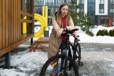 Karlı bir arazide park halindeki bir bisikletin yanında duran bir kadın. Kışlık giysiler giyiyor ve kameraya bakıyor..