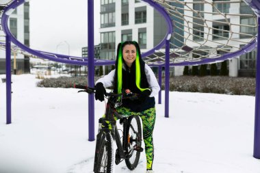 Karlı bir arazide bisikletin yanında duran bir kadın. Kadın soğuk havaya rağmen bisiklete binmeye hazır görünüyor..