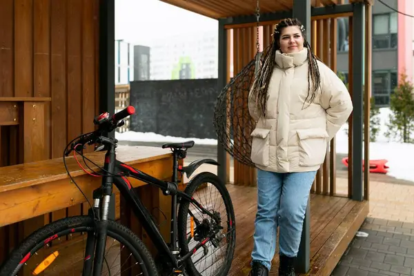 stock image A woman is standing next to a bike on a wooden platform. She appears to be preparing to ride the bike or taking a short break. The wooden platform is surrounded by greenery, creating a natural setting