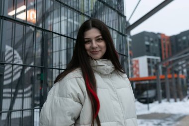 Bir kadın, yüksek bir binanın önünde duruyor. Yapıya kıyasla küçük görünüyor. Bina modern ve parlak, gökyüzüne kadar uzanıyor..