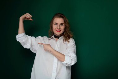 Beyaz gömlek giyen bir kadın fotoğraf için poz veriyor. O, kendinden emin bir ifadeyle doğrudan kameraya bakar. Ayarlar sıradan bir açık hava konumu gibi görünüyor..